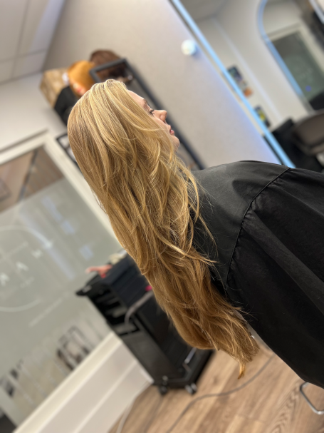 Haircut long layers London Woman with long, wavy blonde hair facing sideways in a salon setting.